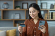 © Liubomir - Asian woman using phone and card for online shopping at home. Relaxed on sofa, she engages in digital transactions, highlighting modern convenience and technology integration in daily life.