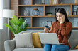 © Liubomir - Asian woman sitting on sofa at home, using laptop for online shopping. Holding credit card with worried expression, symbolizing financial stress and uncertainty.
