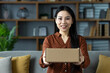 © Liubomir - Smiling woman presenting cardboard box while sitting on sofa in modern home environment. Her expression conveys warmth, inviting experience. Ideal for concepts like delivery, gift, hospitality.
