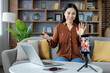 © Liubomir - Asian woman engaging in video conference at home. Sitting on sofa with laptop and phone tripod. Modern living room setting. Concept of remote work, online communication.