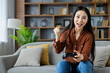 © Liubomir - Asian woman playing video games at home on couch, holding controller with excitement. Joyful gaming experience, expressing thrill and competitive spirit in comfortable living room setting.
