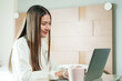 © Ekkasit A Siam - Asian female office worker smiling while working on laptop at home, balancing work and relaxation in a cozy bedroom environment, showcasing modern remote work lifestyle.