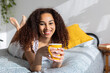 © Xavier Lorenzo - Happy young latin american woman lying on bed, holding a yellow mug and enjoying a relaxing morning with a cup of coffee