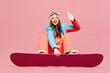 © ViDi Studio - Full body young happy woman wear warm blue padded jacket ski goggles mask spend weekend winter season in mountains sit with snowboard waving hand rest isolated on plain pink background. Hobby concept.