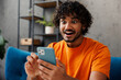 © ViDi Studio - Shocked young Indian man wear orange casual clothes sits on blue sofa couch hold use mobile cell phone stay at home hotel flat rest relax spend free spare time in living room indoor. Lounge concept.