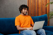 © ViDi Studio - Young IT Indian man wear orange casual clothes sit on blue sofa couch hold use work on laptop pc computer stay at home hotel flat rest relax spend free spare time in living room indoor Lounge concept