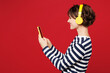 © ViDi Studio - Side view young happy woman she wear blue striped sweatshirt casual clothes listen to music in headphones hold in hand use mobile cell phone isolated on plain red background studio. Lifestyle concept.