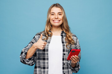 Wall Mural - Young smiling woman wears checkered grey shirt casual clothes hold in hand use mobile cell phone show thumb up isolated on plain pastel light blue cyan background studio portrait. Lifestyle concept.