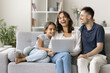© fizkes - Young laughing parents with preschool daughter resting on sofa watching family comedy movie through online digital virtual streaming services using modern laptop, having fun on internet. Tech, leisure