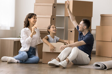 Happy homeowners family take break, resting on laminate floor on move-out or relocation day, enjoy funny playtime with cute little daughter sitting inside big cardboard box. Bank mortgage, new house