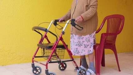 Wall Mural - Elderly woman standing up leaning on walker and starting to walk, sitting on red chair, exercising her legs by walking. Concept of objects to facilitate the daily life of people with disabilities