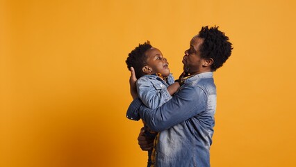 African american dad caressing and kissing his adorable baby boy in studio, carrying his toddler and playing around with him. Cute family showing love and devotion to his infant son. Camera B.