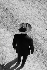 Wall Mural - Man in a black suit and hat stands in front of a rocky hill. The man is a businessman, as he is wearing a suit and hat. He is standing in the middle of the rocky hill, looking up at the sky