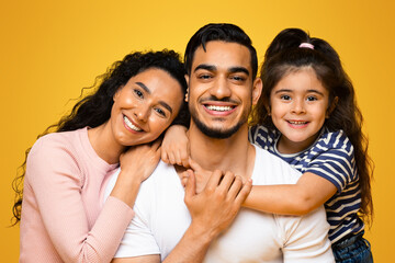 Wall Mural - Closeup Portrait Of Happy Middle Eastern Family Of Three With Adorable Little Daughter, Joyful Middle Eastern Parents And Their Cute Female Child Embracing And Posing At Camera Over Yellow Background