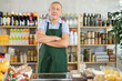 © JackF - Elderly man in green apron stands near counter, works as salesman in grocery store. He is ready to meet customers, offer goods, sell food and drinks.