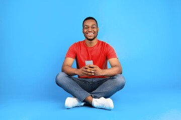 Happy african american male using cellphone smiling to camera sitting on blue studio background. Black guy using new mobile app on smartphone. People and modern gadgets concept