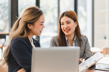 Wall Mural - Two businesswomen in formal attire discussing work at desk with laptop and documents, smiling and engaging in bright office environment