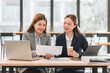 © PRIME STOCK LAB - Two businesswomen in formal attire discussing documents at modern office table with laptops, tablet, and coffee, showcasing teamwork and collaboration in professional setting