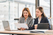© PRIME STOCK LAB - Two businesswomen in formal attire discussing documents at modern office table with laptop, tablet, and coffee cup, showcasing teamwork and collaboration in professional setting