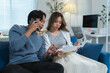 © crizzystudio - Asian couple sitting on sofa at home managing domestic finances, checking bills and bank statements, making phone calls to customer service, using smartphone and laptop