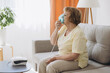 © anatoliycherkas - Sick senior woman has a viral infection or flu, labored breathing, asthma. An elderly female is using inhaler, nebulizer for self treatment sitting alone on the sofa at home