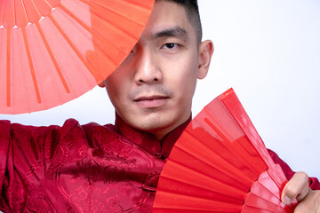 Close-up of an Asian man in a red traditional Chinese Tang suit framed by two red hand fans, symbolizing cultural elegance and mystery, isolated against a plain white background