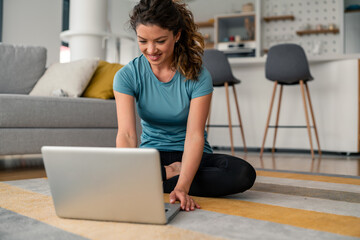 Caucasian woman in casual attire works remotely from home, sitting comfortably on living room floor while using laptop for professional tasks.