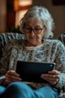 © vefimov - Elderly lady with glasses reading on a digital tablet indoors.