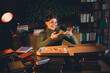 © deagreez - Young woman in a cozy apartment with a green jumper and apron, enjoying a pizza while studying amidst books and a warm evening light