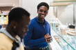 © Studio Marmellata - A cheerful black man in a blue sweater smiles brightly while interacting with a friend in a modern cafe, surrounded by a neatly arranged counter and warm lighting