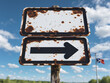 © HooSv - Rusty directional road sign with an arrow, set against a bright blue sky and a distant flag.