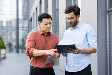 Wall Mural - Two diverse male office workers outside office building discussing documents, company financial report. Colleagues in casual clothes looking at contracts and invoices.