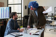 © pressmaster - Male clown boss with pack of paper documents in hand looking at young subordinate while speaking about financial points