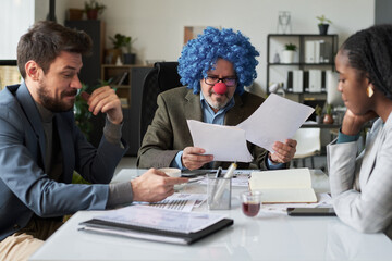 Poster - Mature male director of business company wearing clown wig looking through financial paper documents at meeting with subordinates