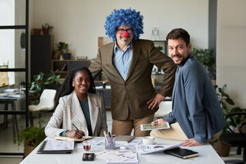 Poster - Two happy young intercultural subordinates surrounding mature clown boss in blue curly wig and red nose while looking at camera