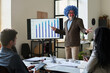 © pressmaster - Happy mature man in formalwear and clown wig standing by interactive board and looking at employees while explaining charts