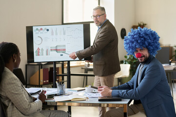 Poster - Young smiling businessman in clown wig looking at camera while sitting by workplace against mature boss making presentation