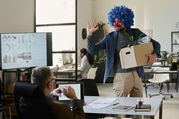 Poster - Happy young clown employee with box waving hand to boss and looking at him while leaving office after being fired