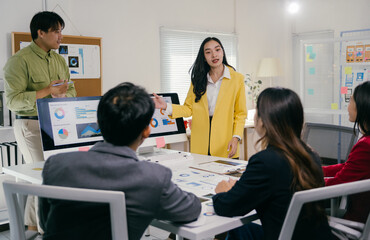 Fototapete - Young businesswoman wearing yellow jacket is leading a business meeting with her colleagues and pointing at charts displayed on computer screen