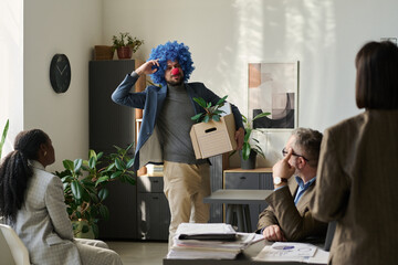 Poster - Young male clown employee with box looking at group of colleagues and keeping fingers by head while leaving office