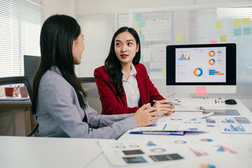 Fototapete - Two asian businesswomen discussing financial charts and data on a computer screen, analyzing company performance and making strategic decisions during an office meeting