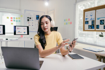 Wall Mural - Asian businesswoman is sitting at her desk wearing headphones and using a smartphone while working on a laptop computer