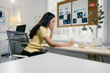 Sticker - Young asian businesswoman concentrating and writing something in notebook while sitting at her desk in modern office with graphs on cork board