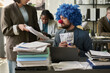 © pressmaster - Young businessman in clown wig and nose looking at stack of paper documents held by female colleague in office