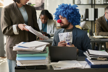 Poster - Young businessman in clown wig and nose looking at stack of paper documents held by female colleague in office