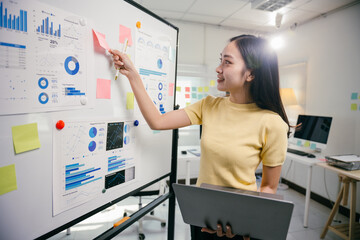 Wall Mural - Young businesswoman is using a laptop and pointing at charts on a whiteboard during a financial data analysis meeting