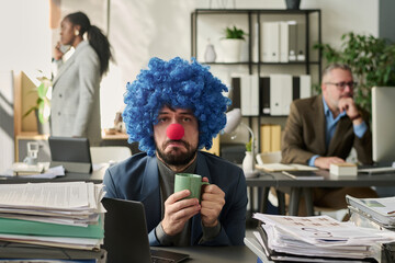 Poster - Young unhappy clown office worker holding cup of tea or coffee and looking at you while sitting by desks with stacks of papers