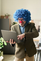 Poster - Happy mature boss in clown wig looking at camera in office while using laptop and pointing at screen during presentation