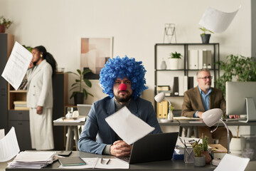 Poster - Young clown male employee with financial paper documents falling around looking at camera while sitting by workplace
