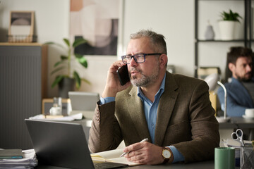 Poster - Confident male director of firm with smartphone by ear organizing work in front of laptop and making notes in notebook